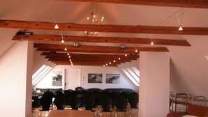 Ein geräumiger Raum mit einem langen Holztisch, mehreren Stühlen, einem braunen Ledersofa und einer hohen Decke mit freiliegenden Holzbalken und Pendelleuchten.