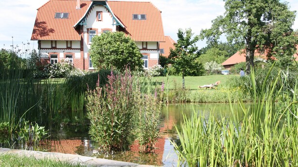 Ein Haus mit rotem Ziegeldach, einem Teich und umgebender Bepflanzung.