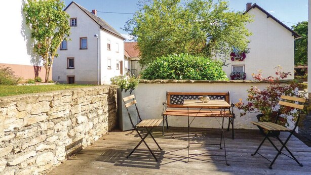 A patio with a wooden table and chairs, a stone wall, and a white building with a sloped roof. - Vacation cottage Ferber (Wiesbaum)