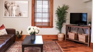 Un salón con un sofá de cuero marrón, una mesa de centro con un florero, un ventanal con persianas, un televisor sobre un mueble de madera y un cuadro en la pared.