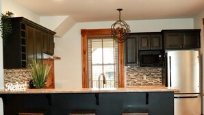 Una cocina con armarios oscuros, un refrigerador de acero inoxidable, protector contra salpicaduras de piedra y una zona de bar con taburetes.
