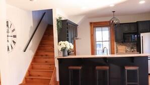 Una cocina con escalera de madera, armarios negros, encimera de mármol y tres taburetes.