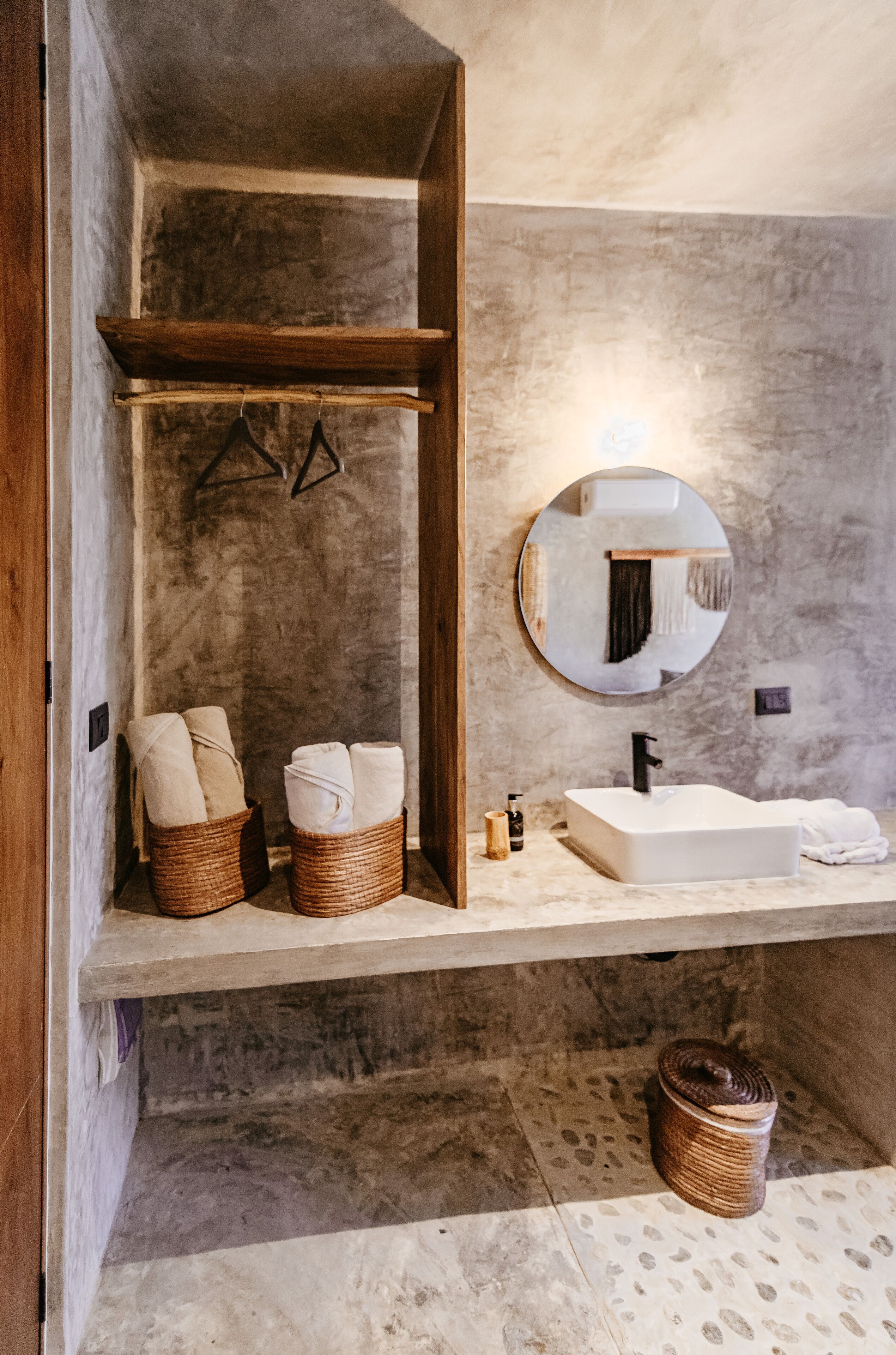 A bathroom with a wooden shelf, a round mirror, a white sink, and two wicker baskets with towels.