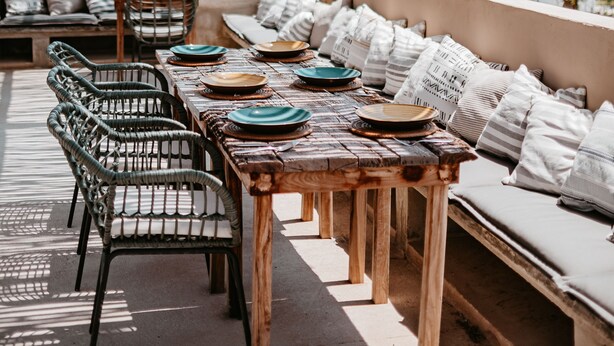 Een eetgedeelte aan het strand met een houten tafel en stoelen, een rieten dak en uitzicht op zee.