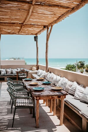 A beachside dining area with a wooden table and chairs, a thatched roof, and a view of the ocean.