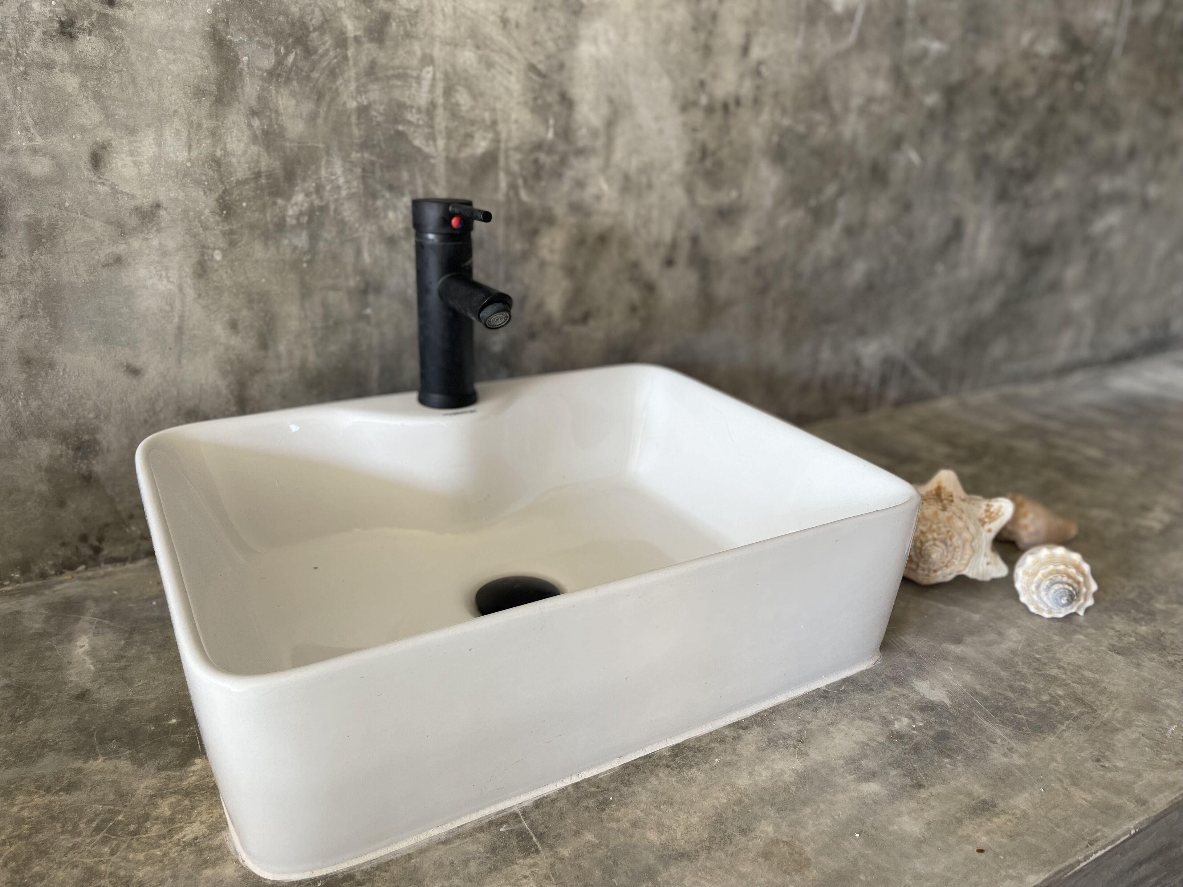 Un évier rectangulaire blanc, avec un robinet noir, adossé à un mur en béton.
