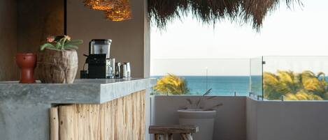 Terraza de madera con barra, incluyendo taburetes y una cafetera, con vistas al mar.