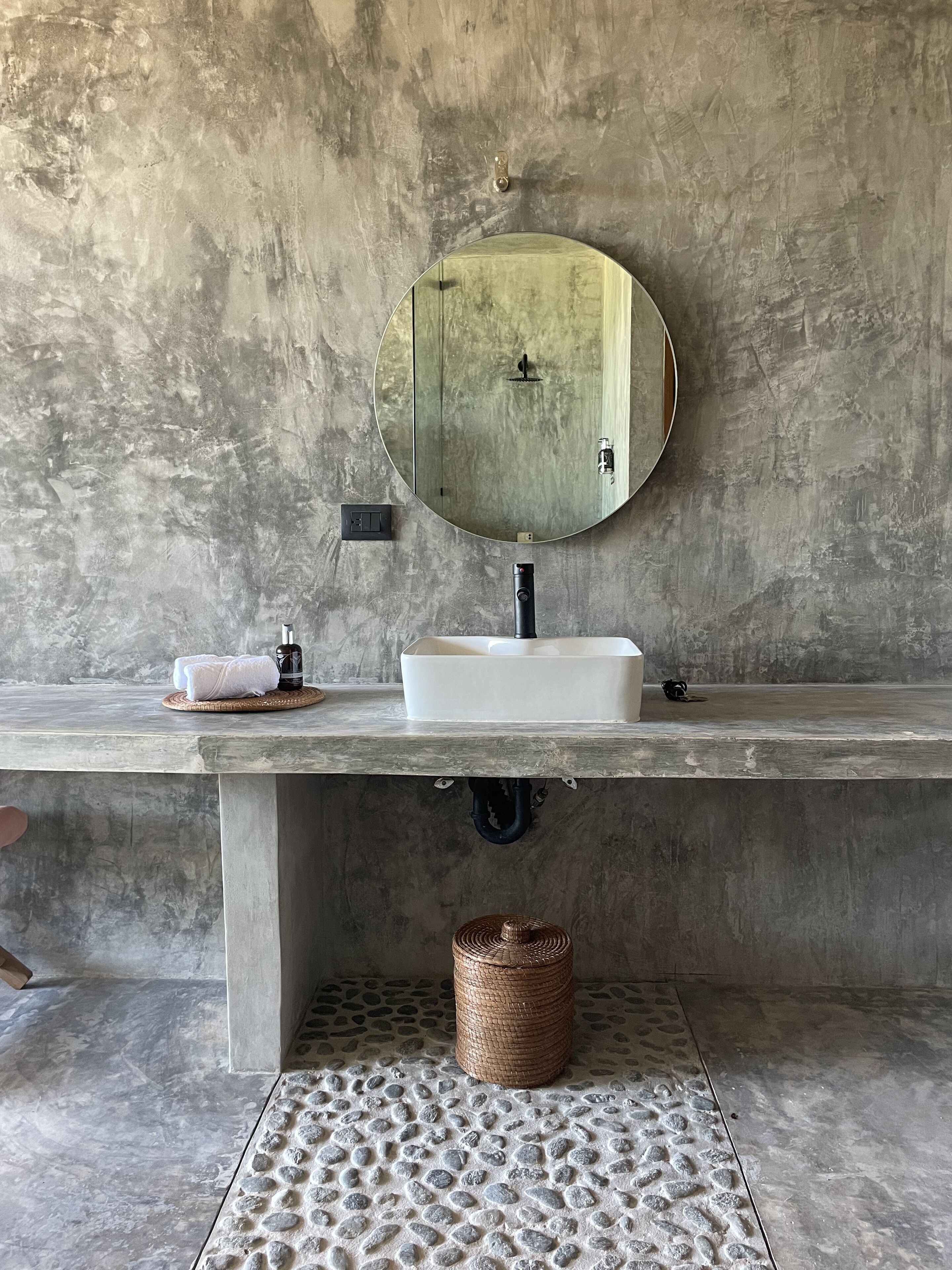 A bathroom with a large, round mirror, a white sink, and a textured stone floor.