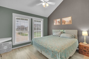 A bedroom with a bed, two windows with blinds, a ceiling fan, a bedside lamp, and a small wooden nightstand.