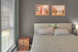 Een slaapkamer met een bed, twee foto's in lijsten aan de muur, een raam met jaloezieën en een houten doos.