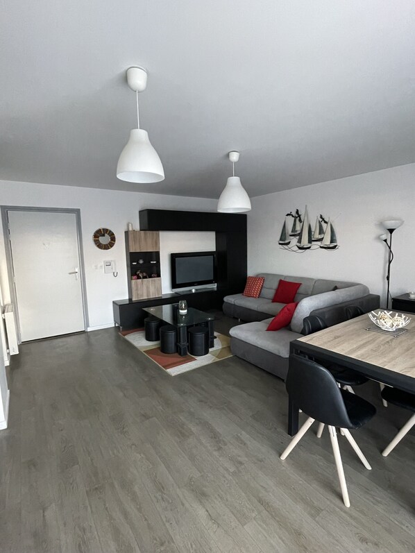 A modern living room with a grey sofa, a dining table, and a TV. There are two pendant lights, a fireplace, and a wall with sailboat decorations. - 100 metres from the beach, great apartment (Lacanau Océan)
