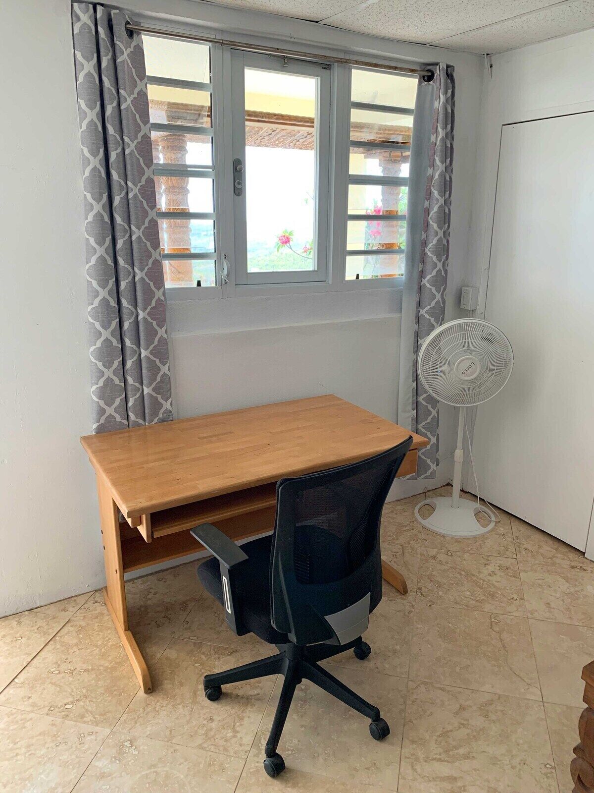 Une pièce avec un bureau en bois, un fauteuil de bureau noir et un ventilateur sur pied.