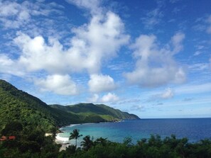A coastal landscape with a beach, lush green hills, and a clear blue sky.
