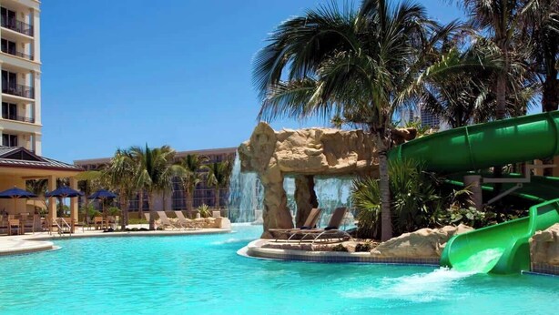 A swimming pool with a water slide, lounge chairs, palm trees, and a building in the background. - The Place (North Palm Beach)
