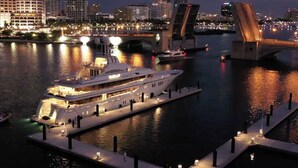 A large, multi-deck yacht docked at a pier with a city skyline in the background. - The Place (North Palm Beach)