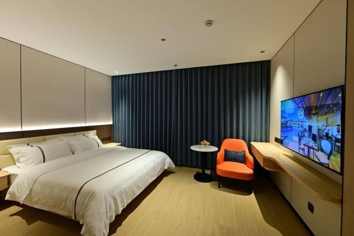 A modern hotel room with a large bed, a small round table, an orange chair, and a large TV mounted on the wall.