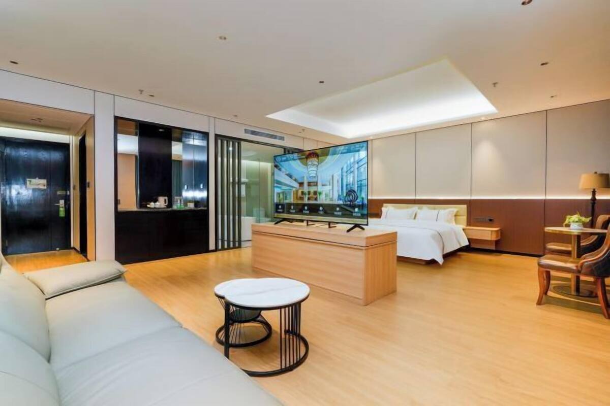 A modern hotel room with a sofa, a small round table, a bed, a TV, and a wooden cabinet.