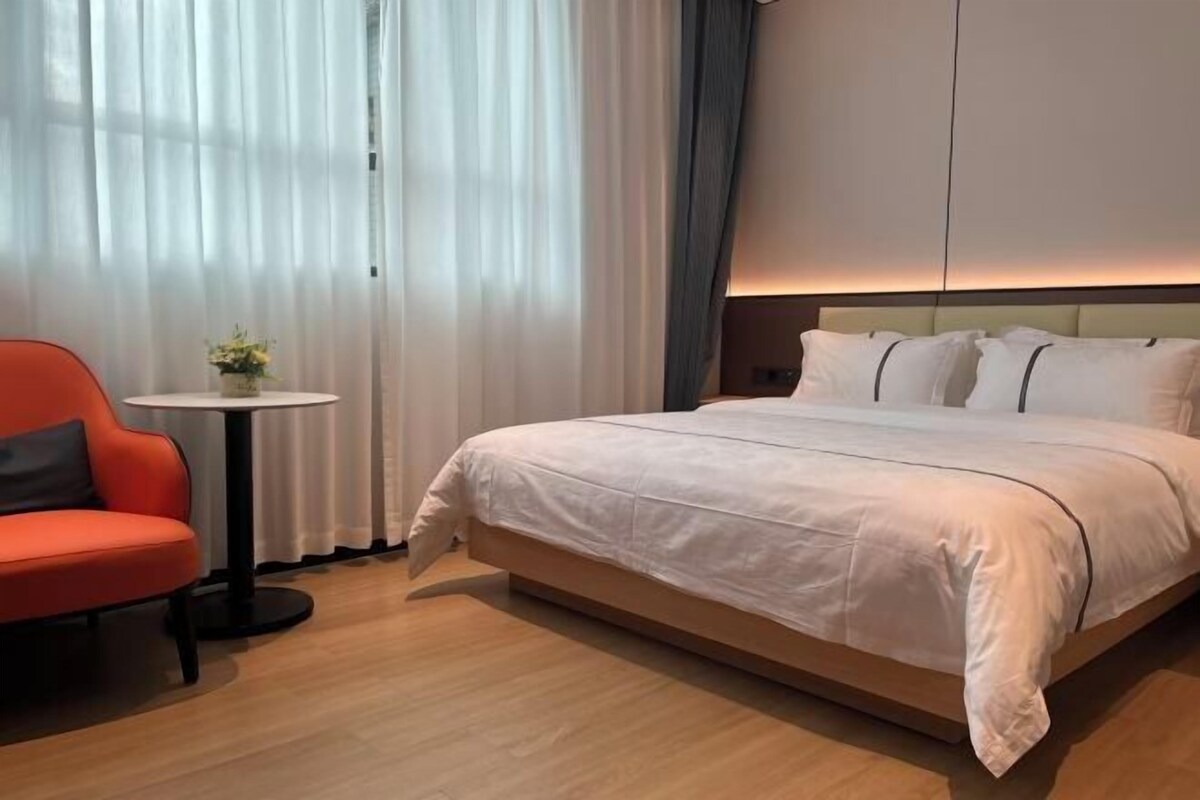 A hotel room with a bed, a small table with a vase, a red chair, and sheer curtains.