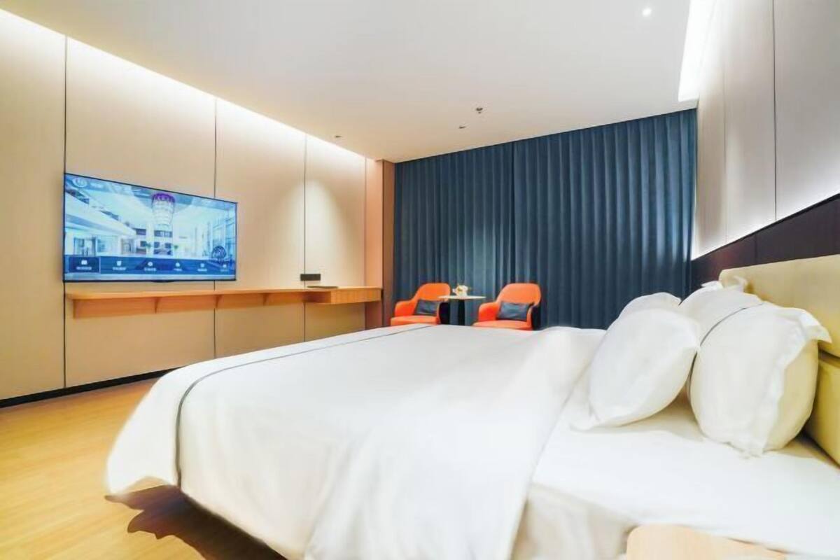 A hotel room with a large bed, a TV mounted on the wall, and two orange chairs.
