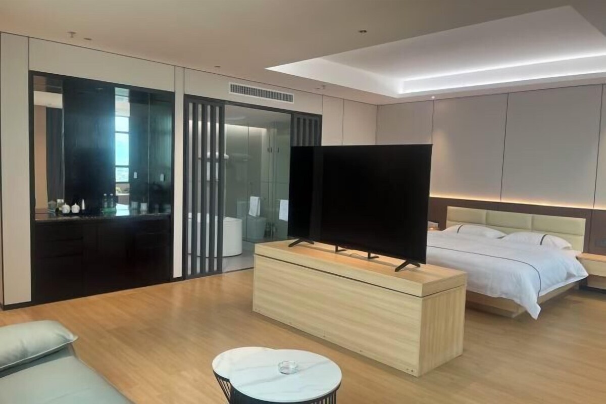 A modern hotel room with a large bed, a TV on a wooden console, a small round table, and a glass-enclosed walk-in closet.