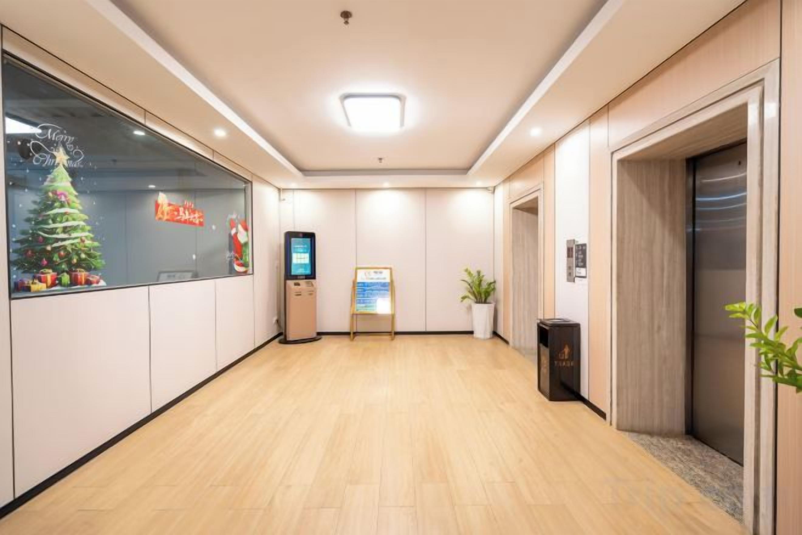 A hotel lobby with a wooden floor, a reception desk, a digital kiosk, a Christmas tree, and an elevator.