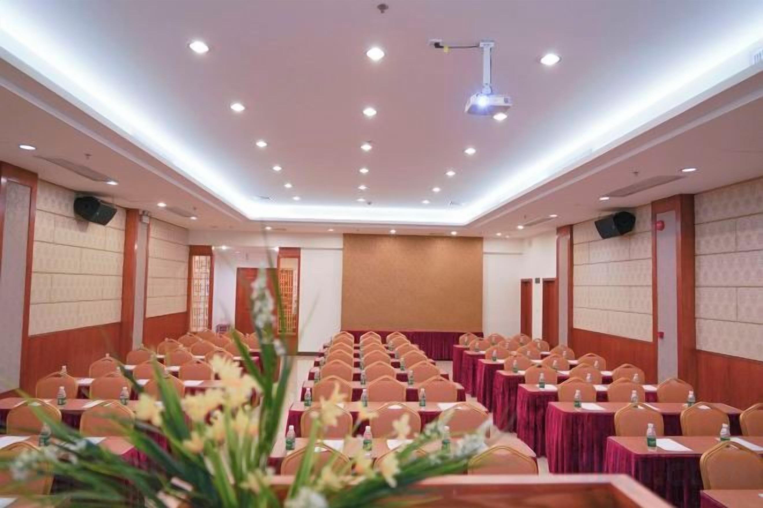 A conference room with rows of tables and chairs, a stage, and a ceiling with recessed lighting.