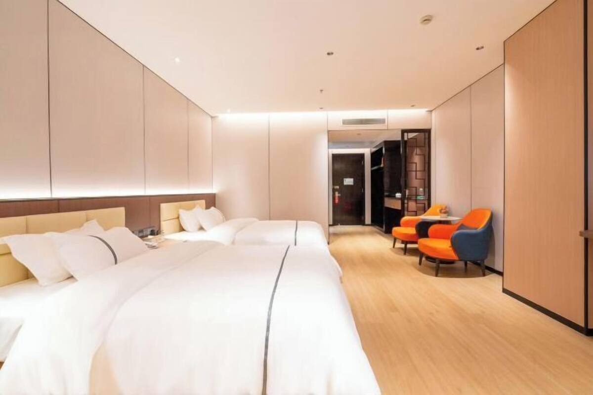 A hotel room with a large bed, a seating area with orange chairs, and a separate room visible through an open door.
