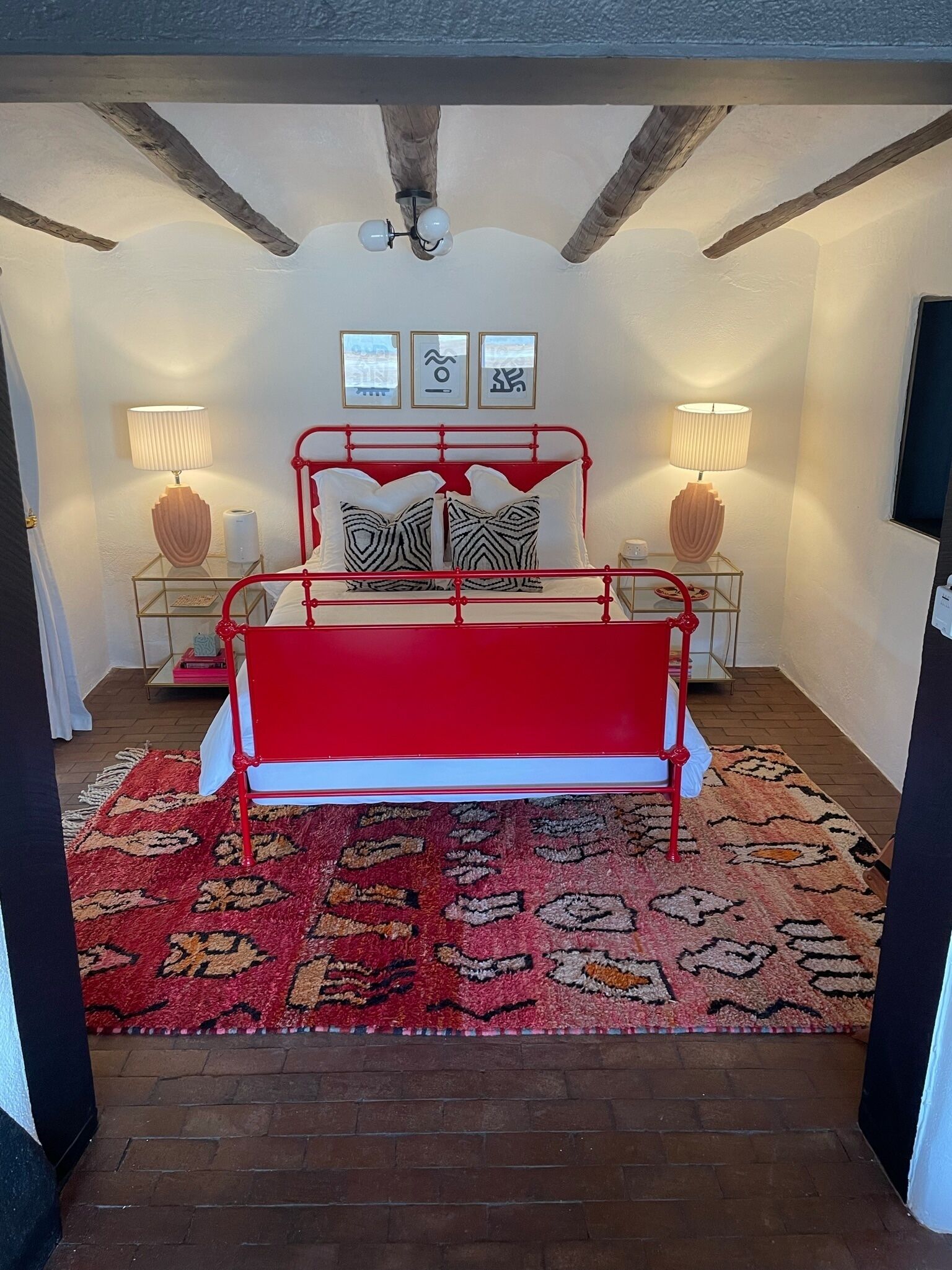 A bedroom with a red metal bed frame, patterned pillows, two bedside lamps, a red and beige rug, and a ceiling fan.