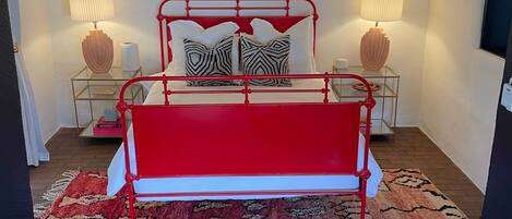 A bedroom with a red metal bed frame, patterned pillows, two bedside lamps, a red and beige rug, and a ceiling fan.