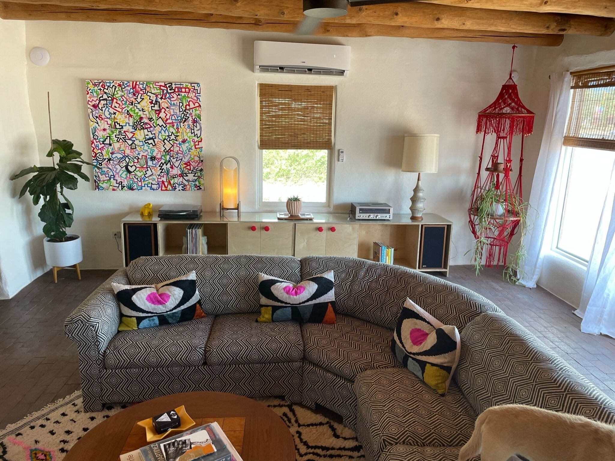 A cozy living room with a patterned sofa, a potted plant, a colorful abstract painting, a lamp, and a red macrame hanging.