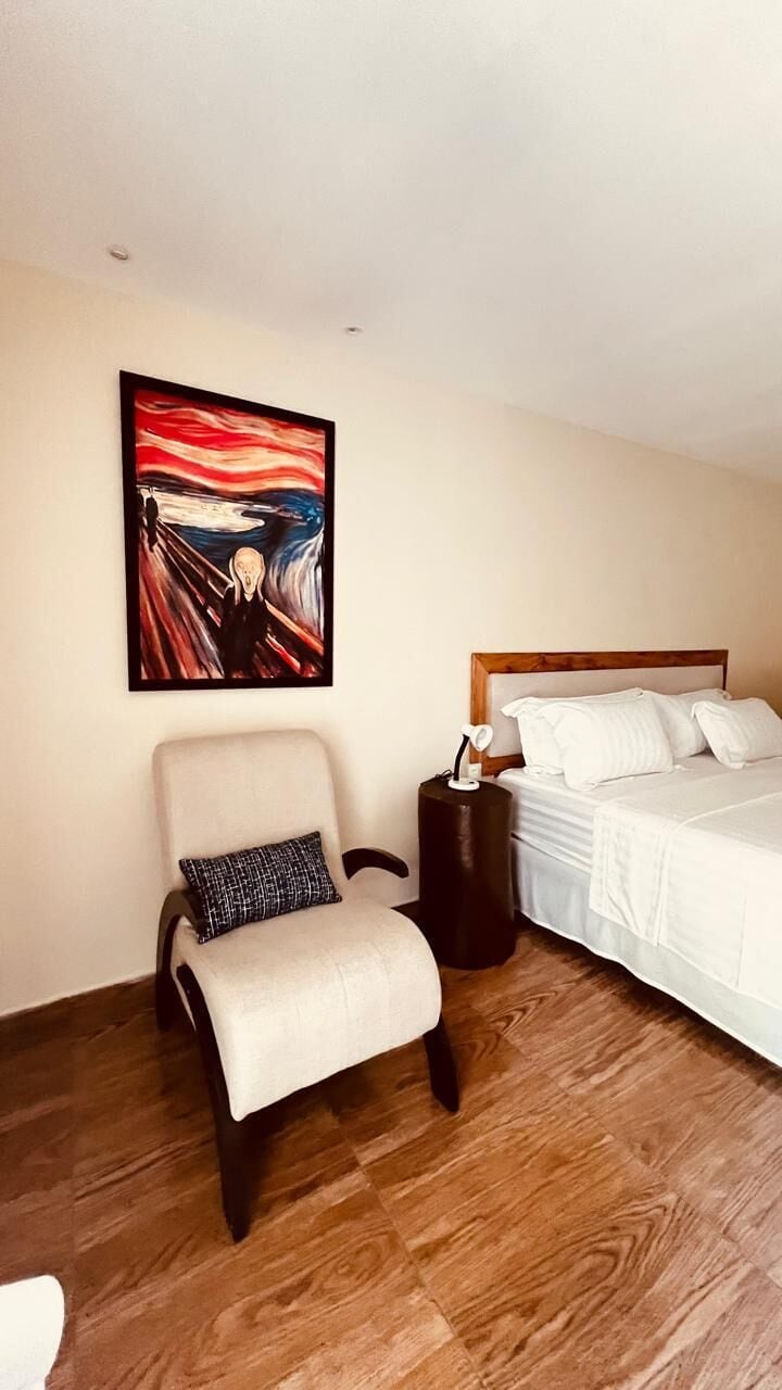 Una habitación de hotel con una cama, una silla, una mesita de noche y un cuadro en la pared.