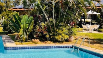 Una piscina azul cristalina rodeada de exuberante vegetación y un área de estar exterior con mobiliario blanco.