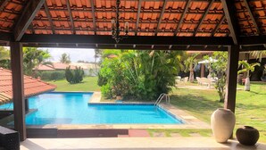 Un área de piscina techada con techo de azulejos, una piscina azul cristalina y abundante vegetación circundante.