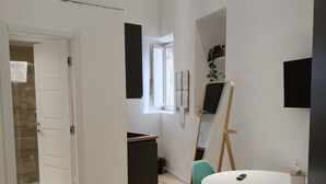 Una habitación moderna con una puerta blanca, una kitchenette pequeña, una mesa redonda con sillas y un espejo grande.