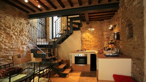 Une cuisine rustique avec un mur en pierre, des poutres apparentes, un four encastré et un coin repas comprenant une table en verre et des chaises en métal.