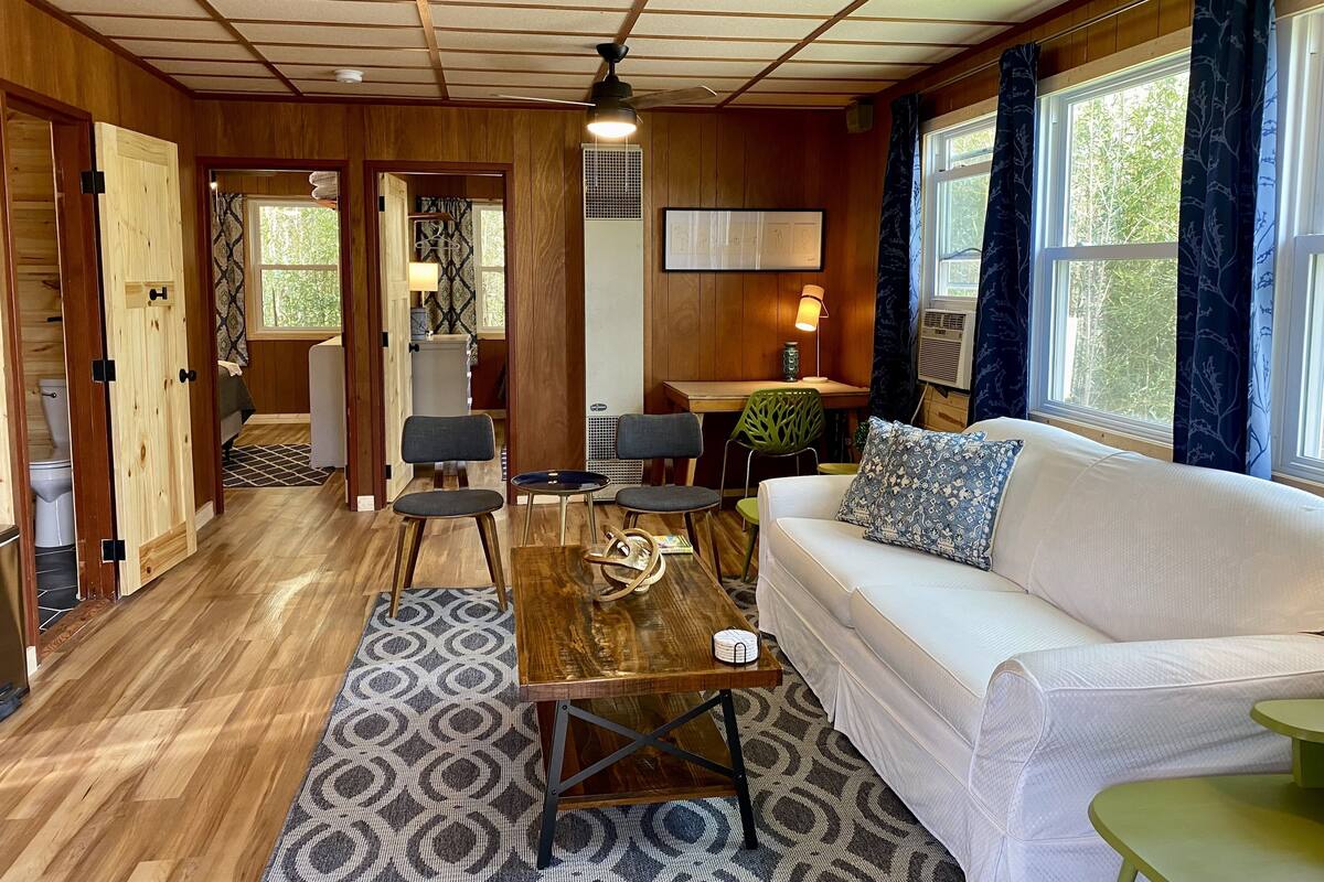 Interior acolhedor de uma cabana com piso de madeira, um sofá branco, uma mesa de jantar e um ventilador de teto.