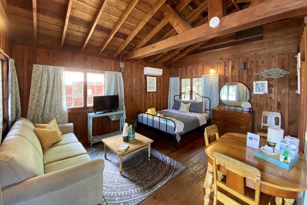 Interior acolhedor de cabana com teto de madeira, sofá, cama, mesa de jantar e televisão.