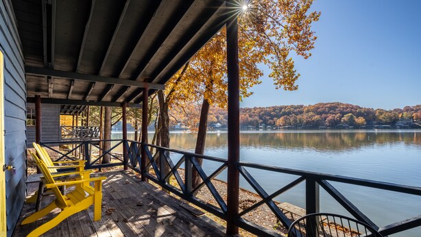A wooden deck with rocking chairs overlooking a calm lake, surrounded by trees with autumn foliage. - Fisherwaters Resort (Camdenton)