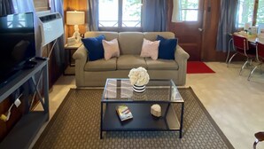 A cozy cabin interior with a wooden ceiling, a sofa with blue and pink cushions, a coffee table with a vase and books, and a television on a stand. - Fisherwaters Resort (Camdenton)