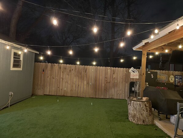 Un patio trasero con césped artificial, cerca de madera, luces colgantes y zona de barbacoa.