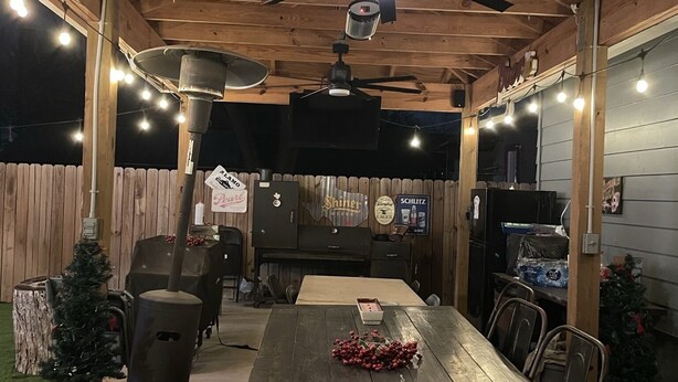 Patio esterno con tavolo in legno, sedie, ventilatore a soffitto, luci a catena e barbecue.