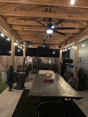Un patio al aire libre con una mesa de madera, sillas, un ventilador de techo, luces colgantes y una parrilla.
