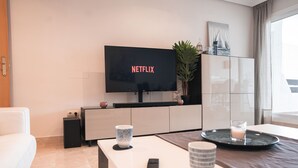 A modern living room with a flat-screen TV displaying 'NETFLIX', a white sofa, a coffee table with two mugs, a remote control, and a decorative plant.