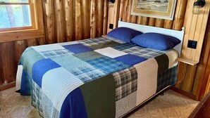 A wooden cabin bedroom with a bed, a window, a wall-mounted picture, and a nightstand.