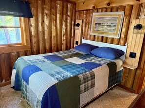 Un dormitori de cabana de fusta amb un llit, una finestra, un quadre penjat a la paret i una tauleta de nit.
