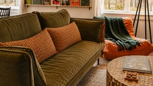 Ein gemütliches Wohnzimmer mit einem grünen Sofa, einem gemusterten Zierkissen, einem runden Couchtisch mit einem Buch und Blick ins Freie durch ein Fenster.