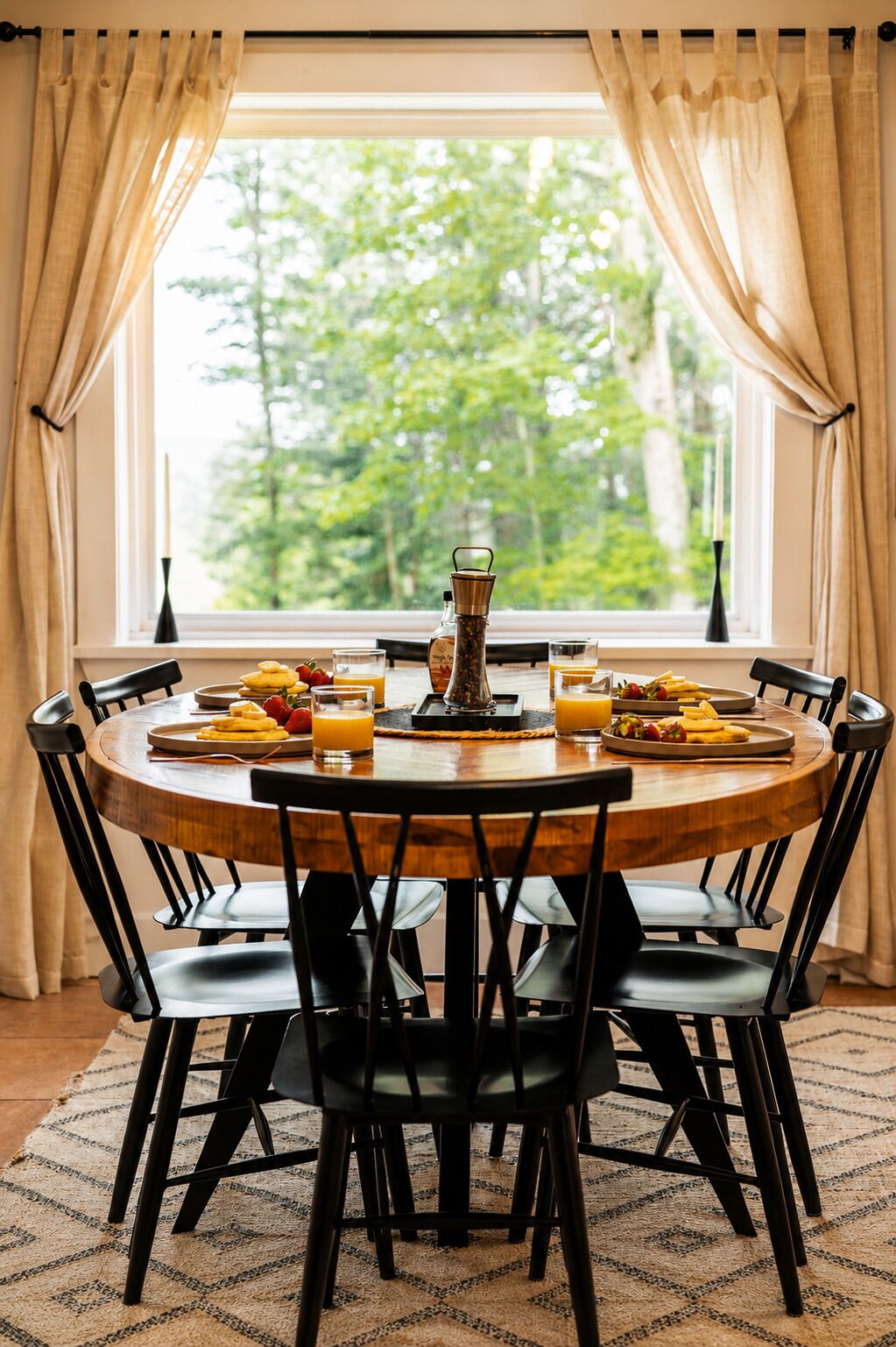 Une table de salle à manger ronde, entourée de chaises noires, est dressée avec des assiettes remplies de nourriture et des boissons, dans une pièce dotée d’une fenêtre et de rideaux.