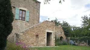 A stone building with a small entrance, a window with shutters, and a garden with flowers and a chair.