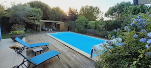 A backyard with a swimming pool, wooden deck, lounge chairs, and a small wooden structure.
