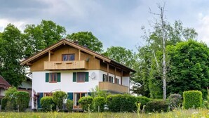 Une maison à deux étages avec un balcon, entourée d’un champ de fleurs sauvages et d’arbres.